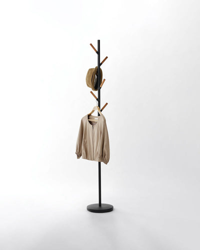 Black Yamazaki Home Coat Rack (70" H) - Steel holding a tan hat and beige jacket on a pale background, with warm brown wood tips contrasting against its dark pole.
