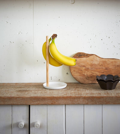 Yamazaki Home Banana Stand - Steel + Wood holding yellow bananas beside wooden cutting boards and a small dark bowl, against a pale speckled kitchen wall.