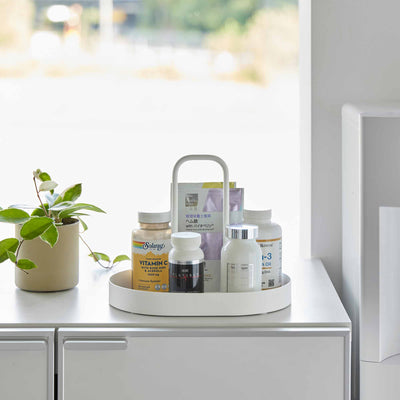Front view of the white Yamazaki Home Vanity Tray with Handle - Steel filled with bottles and packets on a white cabinet beside a leafy plant.