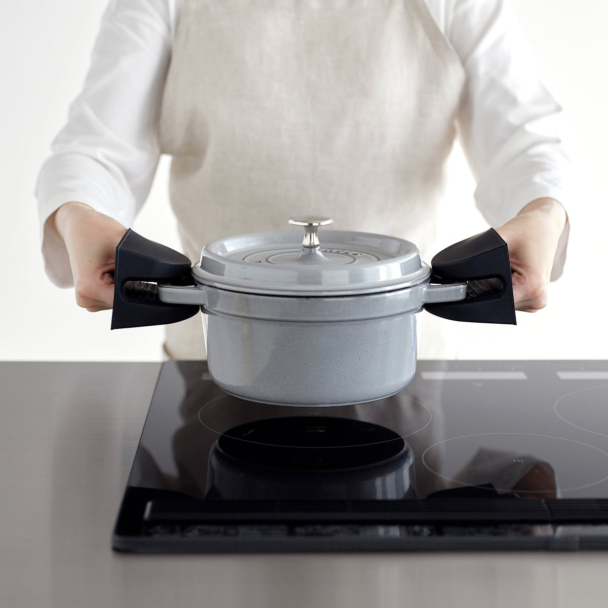 Black Yamazaki Home Magnetic Pot Holders - Set of 2 - Silicone gripping both handles of a grey lidded pot held above a black cooktop by an aproned person.