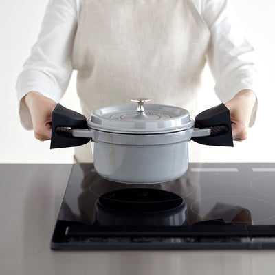 Black Yamazaki Home Magnetic Pot Holders - Set of 2 - Silicone gripping both handles of a grey lidded pot held above a black cooktop by an aproned person.