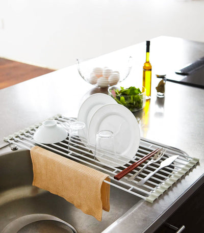 Yamazaki Home Over-the-Sink Dish Drainer - Steel holding upright white plates, a bowl, glass and utensils across a sink, with a mustard-coloured cloth draped over the front edge.