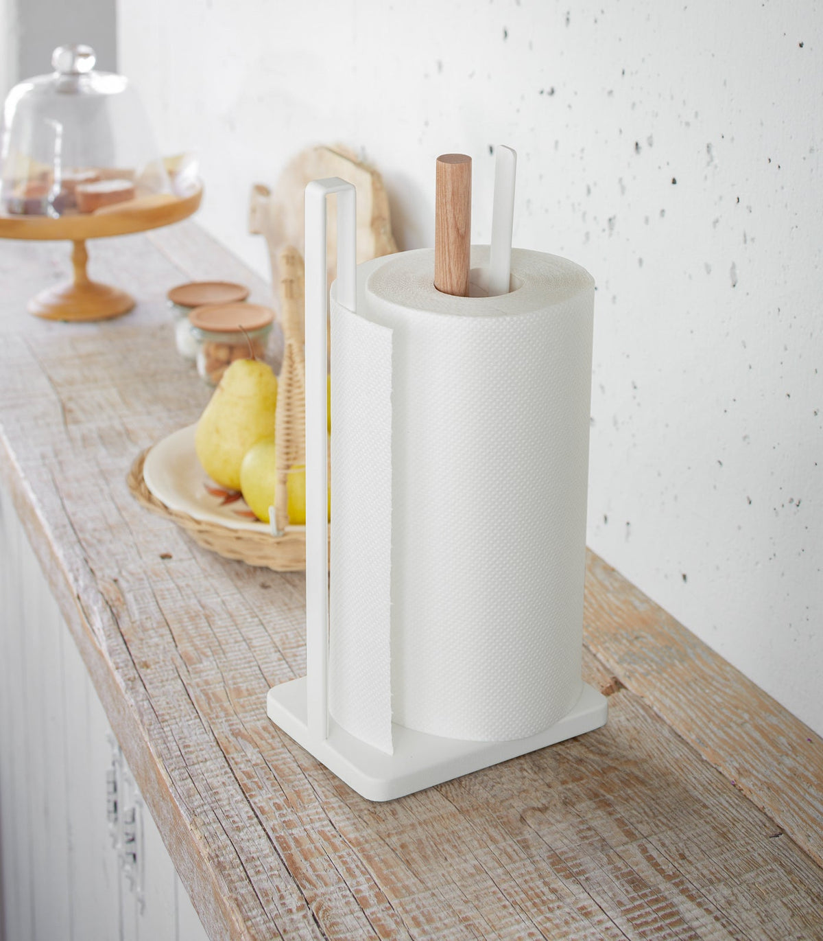 Yamazaki Home Paper Towel Holder - Steel + Wood on a rustic wooden kitchen counter, showing a white frame and natural wood post holding a roll beside lemons and countertop accessories.