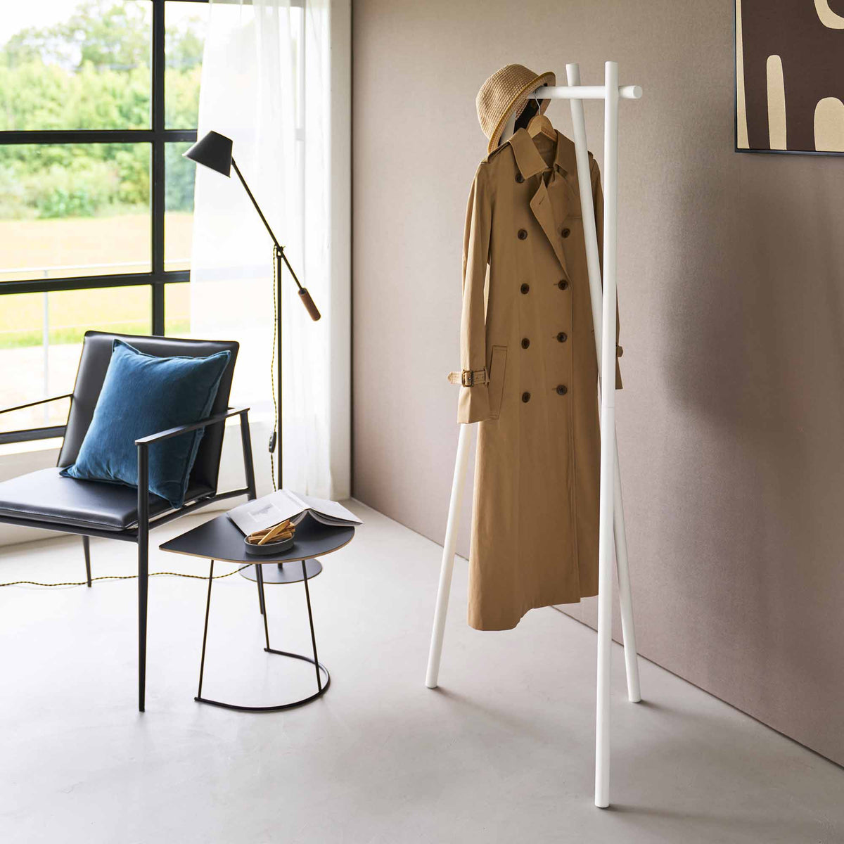 White Yamazaki Home Tri-Post Coat Rack (60" H) - Steel styled with a tan trench coat and straw hat, viewed beside a black chair and small table in a sunlit corner.